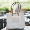 A natural canvas tote bag with natural leather handles, and all of the appellations found within Napa Valley screen printed on the outside. The interior features a wine sleeve to hold wine upright, a pocket, and a clip for keys.